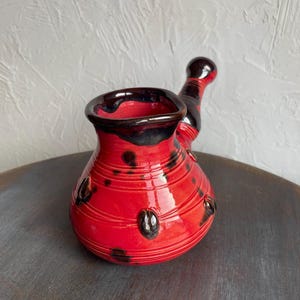 Könnte beinhalten: Eine handgefertigte türkische Kaffeekanne aus roter Keramik mit schwarzem Rand und Griff. Der Topf hat schwarze dekorative Punkte und horizontale Rillen. Der Griff ist ebenfalls schwarz und rot.