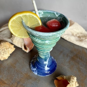 May include: A handcrafted ceramic cocktail glass with a turquoise and blue spiral design. The glass contains red ice cubes, a lemon slice, and a star-patterned straw. Seashells are arranged around the glass on a wooden surface.