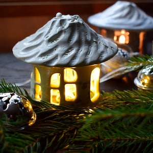 May include: A white ceramic house shaped like a cottage with a lit candle inside, surrounded by green pine branches and silver ornaments.