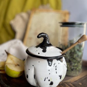 Puede incluir: Tarro de miel de cerámica blanca con un diseño de salpicaduras en blanco y negro. El tarro tiene una tapa con un asa negra en forma de tallo de calabaza. Un cucharón de miel de madera descansa en el borde del tarro.