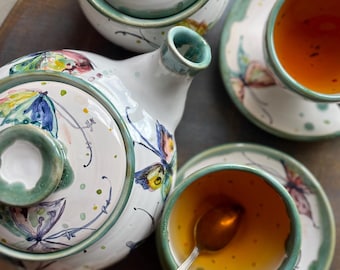 May include: A ceramic tea set with a teapot, sugar bowl, and two teacups with saucers. The set is white with green trim and colorful butterfly and floral designs. The teacups are filled with tea, and one has a silver spoon.