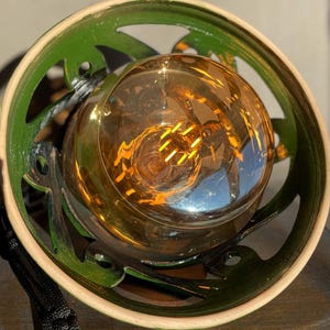 Op de afbeelding: Een groene keramische lampenkap met een uitgesneden ontwerp omringt een heldere glazen gloeilamp met een warme, amberkleurige gloed.