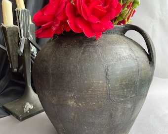May include: A black ceramic vase with two handles, filled with red roses. The vase has a textured surface and is sitting on a white surface.