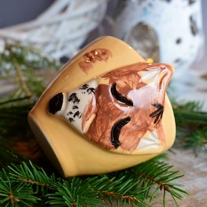 Puede incluir: Una taza de cerámica amarilla con un diseño de zorro pintado a mano. La cara del zorro es marrón y blanca con ojos negros y la boca cerrada. La taza está sobre un lecho de agujas de pino verdes.