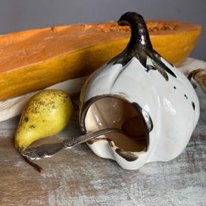 Puede incluir: Un recipiente de cerámica blanco en forma de ajo con un tallo y acentos marrón oscuro, que contiene una cuchara pequeña. También hay una pera amarilla y una sección de una calabaza larga y naranja, lo que sugiere un tema de cocina o comedor.
