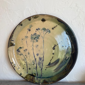 May include: A round ceramic plate with a hand-painted floral design. The plate features a cream-colored center with blue and green floral accents. The rim is a darker green with brown accents. The plate is approximately 12 inches in diameter.
