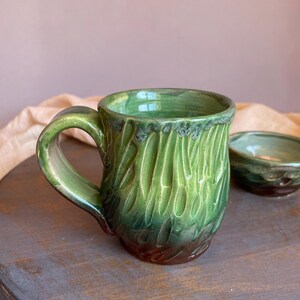 May include: A handmade green ceramic mug with a textured, carved design. The mug has a rounded handle and a dark brown base. A small matching bowl is visible in the background. The mug is approximately 4 inches tall.