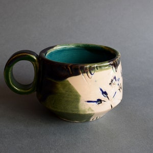 May include: A handmade ceramic mug with a green, blue, and white glaze. The mug has a unique, asymmetrical shape and a wide, rounded handle.
