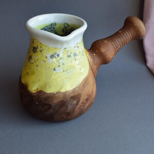 Peut inclure: Une cafetière en céramique avec une base brune arrondie et une partie supérieure texturée émaillée jaune avec des taches grises. Le récipient présente un bord large et évasé avec un bec verseur et une longue poignée brune nervurée.