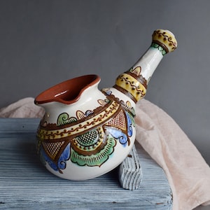 Puede incluir: Una jarra de cerámica blanca con un largo pico curvo. La jarra está decorada con coloridos patrones florales y geométricos en tonos azules, verdes, amarillos y marrones.