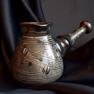 Puede incluir: Una cafetera de cerámica gris con un asa larga y curva. La cafetera tiene una superficie texturizada con un diseño de hojas y líneas. La cafetera está sobre un fondo de tela negra.