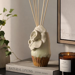 May include: A ceramic essential oil diffuser with a floral design, featuring wooden reeds. The diffuser has a light green and brown color scheme. A lit sandalwood scented candle and a book titled "NATURAL LIVING" are also visible.
