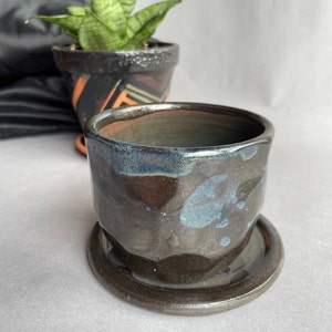 Puede incluir: Un macetero de cerámica negra con un esmalte azul y un platillo a juego. El macetero tiene una superficie texturizada y desigual.