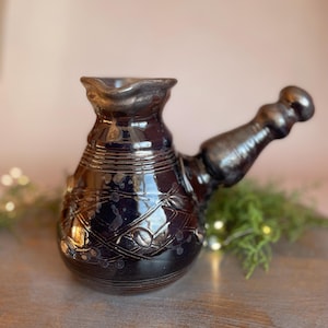 May include: A dark purple and black ceramic coffee pot with a wide base, a narrow neck, and a side handle. The surface features a glossy finish with a decorative pattern of intersecting lines and small circular accents around the body.