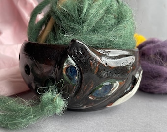 Op de afbeelding: Een zwarte keramische garenkom in de vorm van een kattenkop met blauwe ogen. De kom is gevuld met groen garen en houten breinaalden.