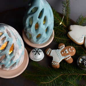 Puede incluir: Una composición festiva con candelabros de cerámica turquesa y blancos con diseños calados, exhibidos sobre platillos rosas. También hay galletas de jengibre y campanas plateadas con diseños de copos de nieve, junto con ramitas de hoja perenne.