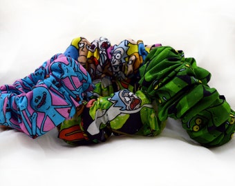 May include: A collection of colorful scrunchies featuring cartoon characters. The scrunchies display various designs, including blue with pink figures, green with white and black figures, and yellow with blue figures. The fabric is gathered, creating a textured appearance.