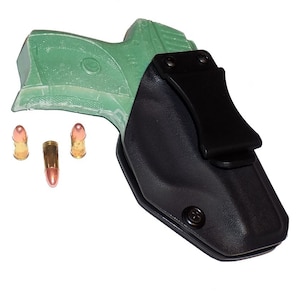 Puede incluir: Funda negra con clip, que sostiene una pistola verde. Tres balas se colocan cerca del arma. Las balas tienen una carcasa de color cobre y una punta plateada. El fondo es blanco.