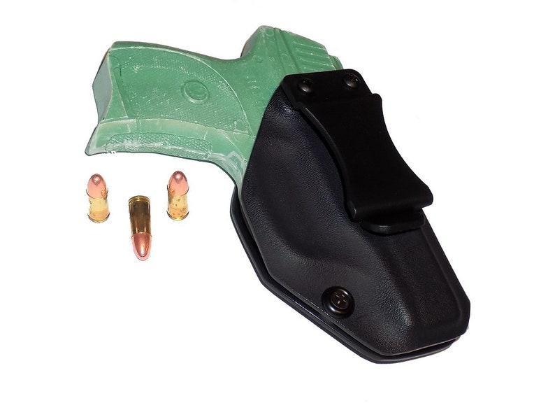 Puede incluir: Funda negra con clip, que sostiene una pistola verde. Tres balas se colocan cerca del arma. Las balas tienen una carcasa de color cobre y una punta plateada. El fondo es blanco.