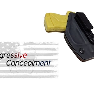 May include: A black holster with a yellow gun inside. The holster has a clip on the back and a textured surface. The text "Aggressive Concealment" is visible in the background.