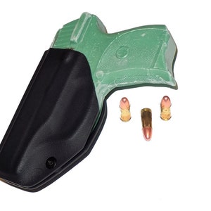 Puede incluir: Una funda negra con una pistola verde en su interior, acompa&ntilde;ada de tres balas con puntas color cobre. Los objetos se presentan sobre un fondo blanco, resaltando el contraste entre la funda oscura y el fondo brillante.