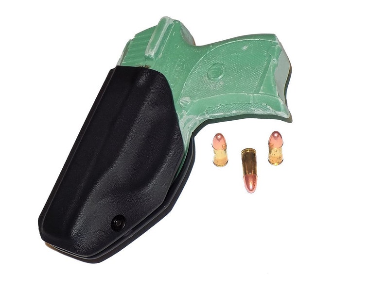 Puede incluir: Una funda negra con una pistola verde en su interior, acompa&ntilde;ada de tres balas con puntas color cobre. Los objetos se presentan sobre un fondo blanco, resaltando el contraste entre la funda oscura y el fondo brillante.