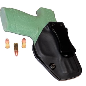 May include: Black leather holster with a black clip, holding a green pistol. Three bullets are placed near the pistol. The holster is designed to be worn on the body.