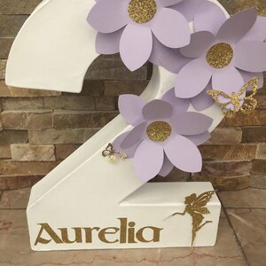May include: A white cardboard number two decorated with lavender paper flowers, gold glitter accents, and gold glitter butterflies. The number has the name "Aurelia" written in gold glitter letters at the bottom.