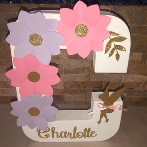 May include: White letter C decorated with pink and purple paper flowers with gold glitter centers. The letter has a silhouette of a fairy with gold glitter and a pink ribbon. The name Charlotte is written in gold glitter below the letter.