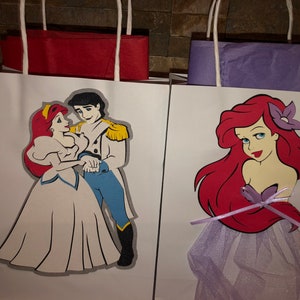 May include: Two white paper gift bags with handles. The bag on the left has a red and white illustration of Ariel and Prince Eric from The Little Mermaid. The bag on the right has a red and purple illustration of Ariel from The Little Mermaid.