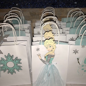 Może przedstawiać: Grupa białych papierowych toreb na prezenty z niebieskimi i srebrnymi wzorami śnieżynek. Niekt&oacute;re torby mają wycięte zdjęcie Elsy z Krainy Lodu, podczas gdy inne mają wycięte zdjęcie Olafa.