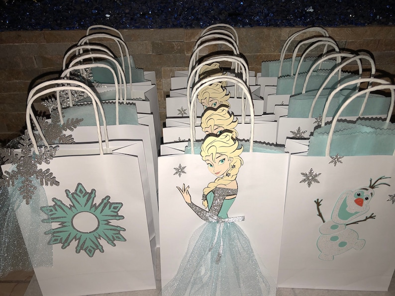 Może przedstawiać: Grupa białych papierowych toreb na prezenty z niebieskimi i srebrnymi wzorami śnieżynek. Niekt&oacute;re torby mają wycięte zdjęcie Elsy z Krainy Lodu, podczas gdy inne mają wycięte zdjęcie Olafa.