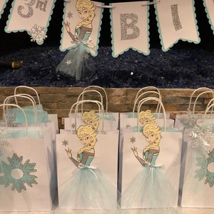 Może przedstawiać: Grupa białych papierowych toreb na prezenty z niebieskimi i srebrnymi wzorami śnieżynek. Każda torba zawiera nadrukowany obraz kresk&oacute;wkowej księżniczki o blond włosach w niebieskiej sukience i białej sp&oacute;dnicy tiulowej. Torby mają białe uchwyty i są ozdobione niebieskimi i srebrnymi błyskotkami.