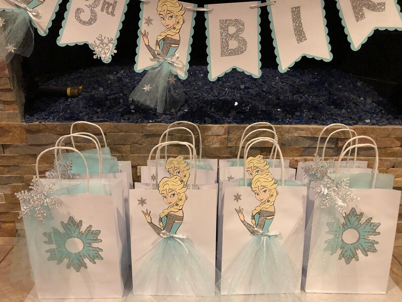 Może przedstawiać: Grupa białych papierowych toreb na prezenty z niebieskimi i srebrnymi wzorami śnieżynek. Każda torba zawiera nadrukowany obraz kresk&oacute;wkowej księżniczki o blond włosach w niebieskiej sukience i białej sp&oacute;dnicy tiulowej. Torby mają białe uchwyty i są ozdobione niebieskimi i srebrnymi błyskotkami.