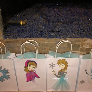 Może przedstawiać: Cztery białe papierowe torby prezentowe z niebieskimi i srebrnymi wzorami śnieżynek. Torby przedstawiają obrazy postaci z filmu Kraina Lodu, w tym Elsę, Annę i Olafa.