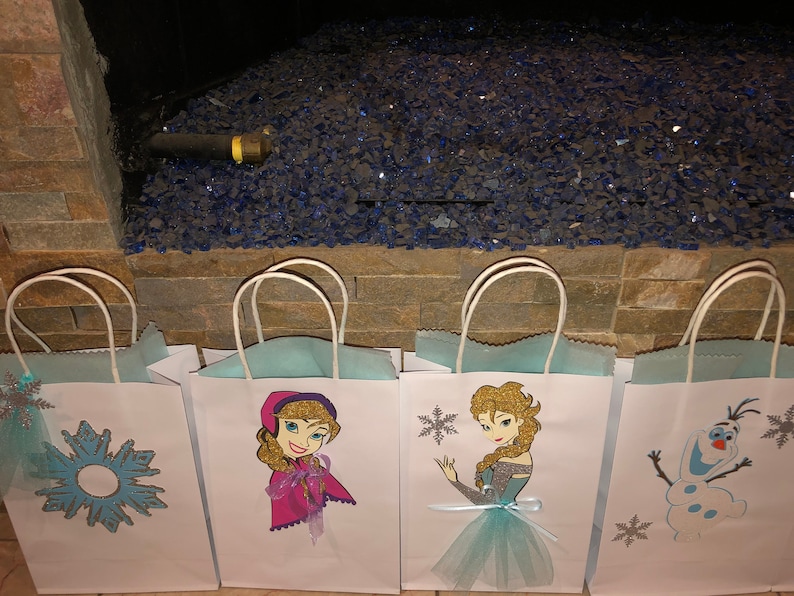 Może przedstawiać: Cztery białe papierowe torby prezentowe z niebieskimi i srebrnymi wzorami śnieżynek. Torby przedstawiają obrazy postaci z filmu Kraina Lodu, w tym Elsę, Annę i Olafa.