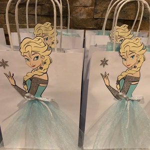 Może przedstawiać: Białe papierowe torby prezentowe z niebieską i srebrną tiulową sp&oacute;dnicą z brokatem i nadrukowanym obrazkiem Elsy z Krainy Lodu. Każda torba ma biały uchwyt.