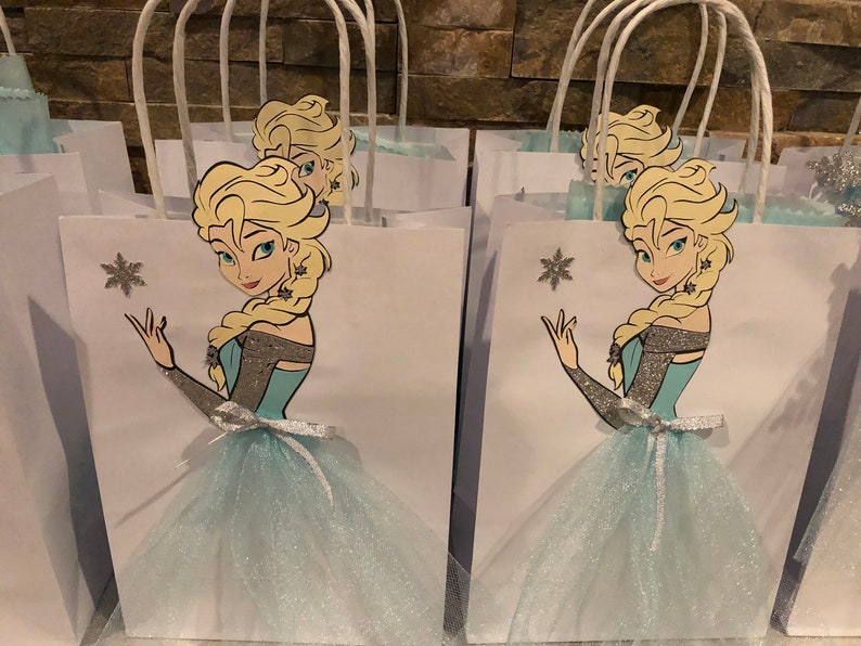 Może przedstawiać: Białe papierowe torby prezentowe z niebieską i srebrną tiulową sp&oacute;dnicą z brokatem i nadrukowanym obrazkiem Elsy z Krainy Lodu. Każda torba ma biały uchwyt.