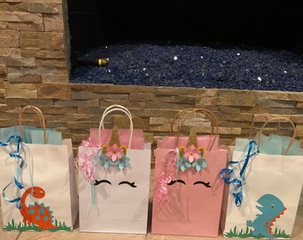 May include: Four gift bags with unicorn and dinosaur themes. Two bags are white with blue and pink accents and feature a unicorn face with a gold horn and flowers. The other two bags are pink and blue with a dinosaur design.