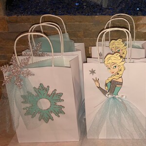 Może przedstawiać: Białe papierowe torebki prezentowe ozdobione wzorem niebieskich i srebrnych płatk&oacute;w śniegu oraz postacią z filmu Kraina Lodu. Postać ma na sobie niebieską sukienkę i białą tiulową sp&oacute;dniczkę.