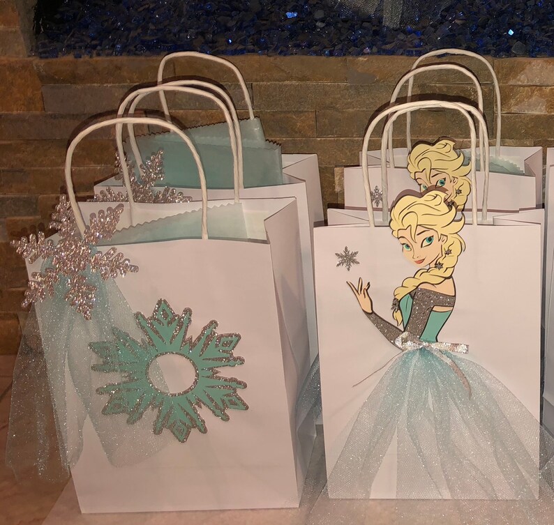 Może przedstawiać: Białe papierowe torebki prezentowe ozdobione wzorem niebieskich i srebrnych płatk&oacute;w śniegu oraz postacią z filmu Kraina Lodu. Postać ma na sobie niebieską sukienkę i białą tiulową sp&oacute;dniczkę.