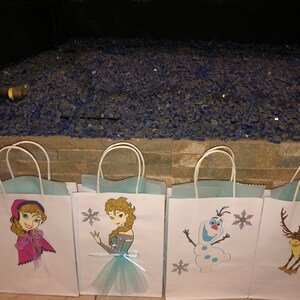 Może przedstawiać: Pięć białych papierowych toreb na prezenty z niebieskimi uchwytami. Każda torba ma inny nadruk z postacią z Krainy Lodu: Annę, Elsę, Olafa i renifera. Torby są wyłożone jasnoniebieskim papierem.