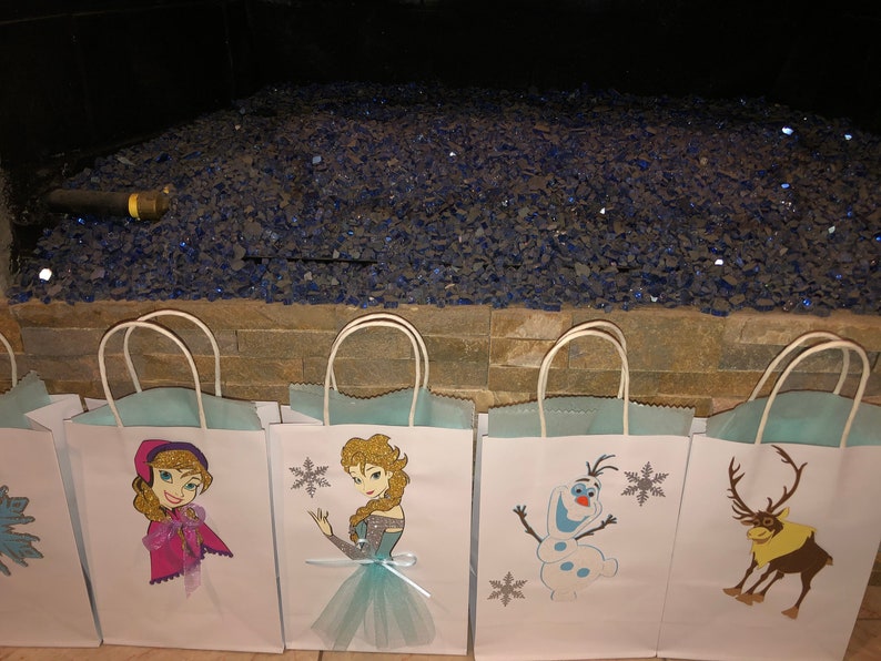 Może przedstawiać: Pięć białych papierowych toreb na prezenty z niebieskimi uchwytami. Każda torba ma inny nadruk z postacią z Krainy Lodu: Annę, Elsę, Olafa i renifera. Torby są wyłożone jasnoniebieskim papierem.