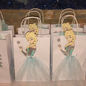 Może przedstawiać: Zestaw białych papierowych toreb na prezenty z wzorem niebieskich i srebrnych płatk&oacute;w śniegu. Każda torba zawiera wycięcie postaci blondynki księżniczki w niebieskiej sukience trzymającej płatek śniegu. Torby mają białe uchwyty i są ozdobione niebieskim tiulem.