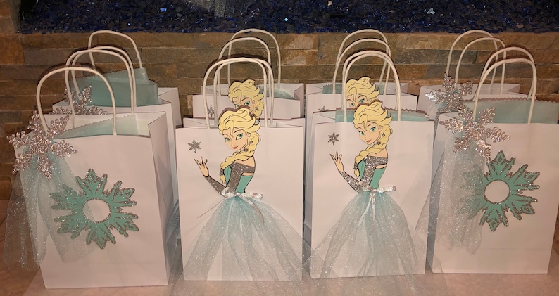Może przedstawiać: Zestaw białych papierowych toreb na prezenty z wzorem niebieskich i srebrnych płatk&oacute;w śniegu. Każda torba zawiera wycięcie postaci blondynki księżniczki w niebieskiej sukience trzymającej płatek śniegu. Torby mają białe uchwyty i są ozdobione niebieskim tiulem.