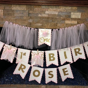 May include: Pink and gold glitter banner with the words "Happy 1st Birthday Rose". The banner is strung with pink tulle.