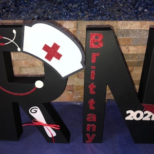 May include: Black foam board letters spelling "RN" with red glitter accents. The "R" has a white nurse's cap with a red cross, a red stethoscope, and a white scroll. The "N" has the name "Brittany" and the year "2021" with a red graduation cap.
