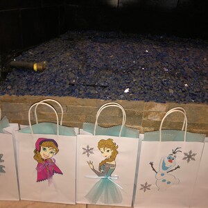 Może przedstawiać: Pięć białych papierowych toreb na prezenty z niebieskimi i srebrnymi wzorami płatk&oacute;w śniegu. Torby przedstawiają obrazy postaci z filmu Kraina Lodu, w tym Elsę, Annę, Olafa i Svena.