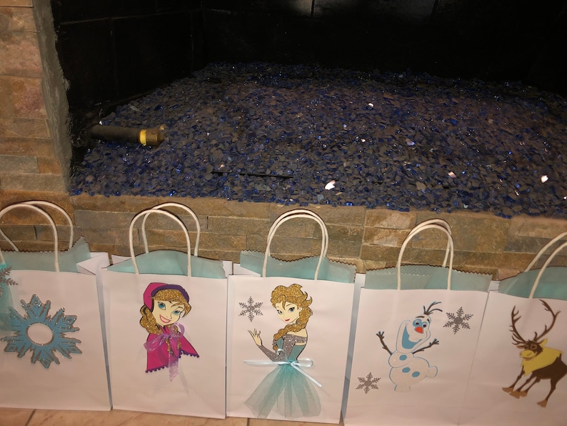 Może przedstawiać: Pięć białych papierowych toreb na prezenty z niebieskimi i srebrnymi wzorami płatk&oacute;w śniegu. Torby przedstawiają obrazy postaci z filmu Kraina Lodu, w tym Elsę, Annę, Olafa i Svena.