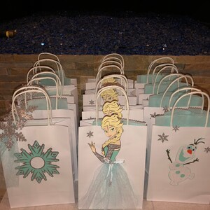 Może przedstawiać: Zbi&oacute;r białych papierowych toreb na prezenty z niebieskimi i srebrnymi wzorami śnieżynek. Niekt&oacute;re torby przedstawiają kresk&oacute;wkową postać Elsy z Krainy Lodu, a inne Olafa.